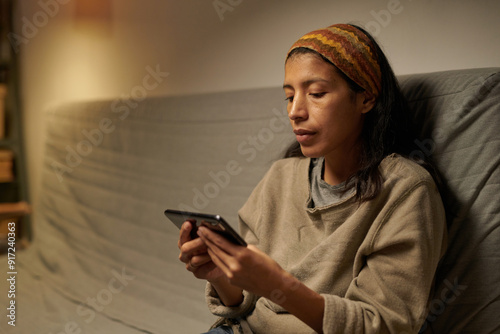 Hispanic woman sitting on sofa using smartphone while surrounded by dimly lit cozy room, wearing casual clothes and focused on screen photo