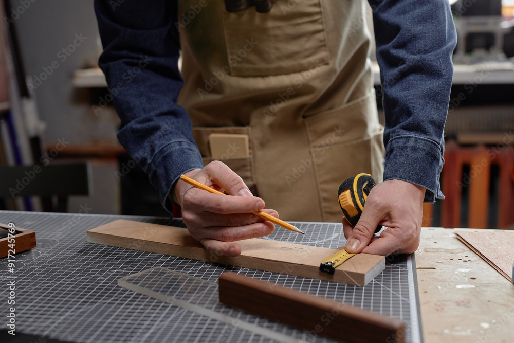 Hands creating detailed measurement on wooden plank using pencil and tool. Person wearing apron and blue shirt in woodworking workshop focusing on perfecting design Hands creating detailed measurement on wooden plank using pencil and tool. Person wearing apron and blue shirt in woodworking workshop focusing on perfecting design