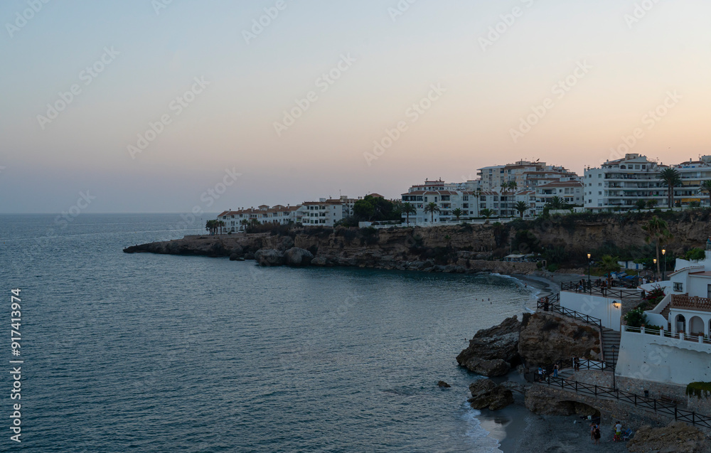 Fototapeta premium A tranquil dusk view of Nerja's coastline, featuring cliffside buildings and the calm Mediterranean Sea. Perfect for travel, sunset, and coastal themes highlighting Spain's beauty.