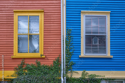 Two exterior colorful houses with vintage closed glass windows. The red building has yellow trim. The blue structure has cream colored trim. The two neighbors have green ivy growing on the clapboard.