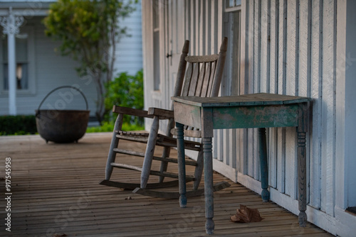 A small wooden accessory table with a vintage wood rocking chair. The furniture is on the back porch of the house. The wooden deck is brown and the house is white. There's a large cast iron cauldron.