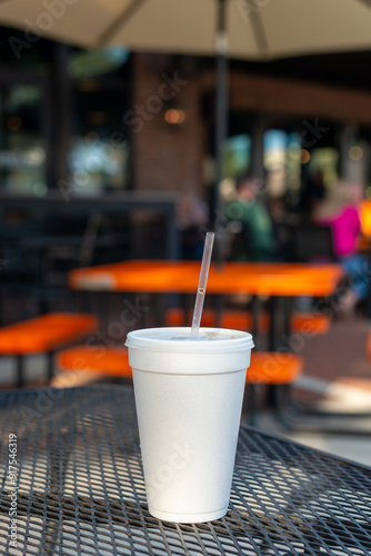 A large white nonrecyclable to-go drink cup with a plastic lid and straw on a black metal patio table. The polystyrene or Styrofoam insulated cup is filled with a cold soda pop drink.  photo