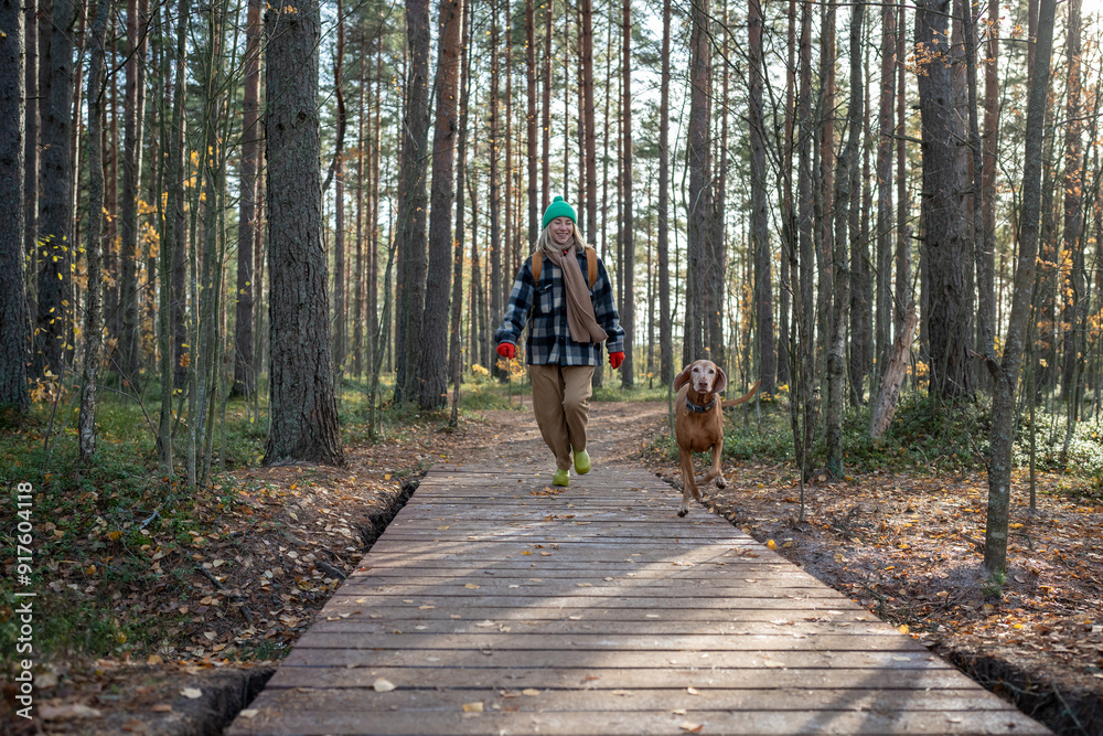 Fototapeta premium Smiling girl with cheerful smile wandering through thicket with running playful beloved pet. Joyful female backpacker together with puppy on hike, traveling, enjoying nature in joint adventure journey
