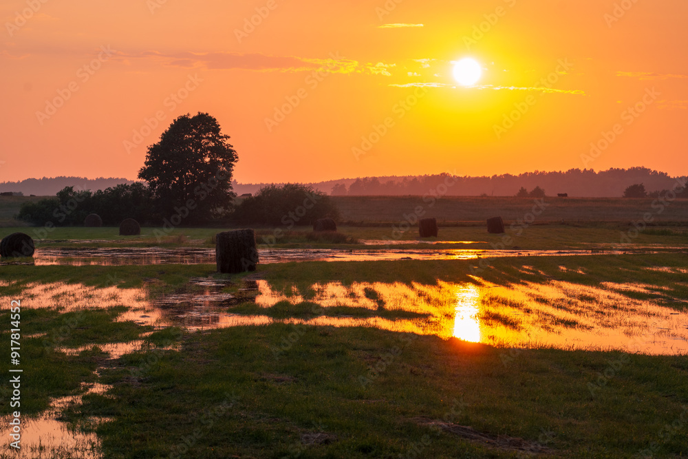 Beautiful rural landscape at sunset moments after a storm. Flooded meadows with hay bales. The setting sun reflecting in puddles
