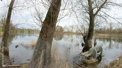 Bench at edge of flooded lake between bare trees, misty background, cloudy winter day in nature reserve Negenoord-Kerkeweerd at De Wissen Maasvallei in Dilsen-Stokkem, Belgium. Pan shot photo