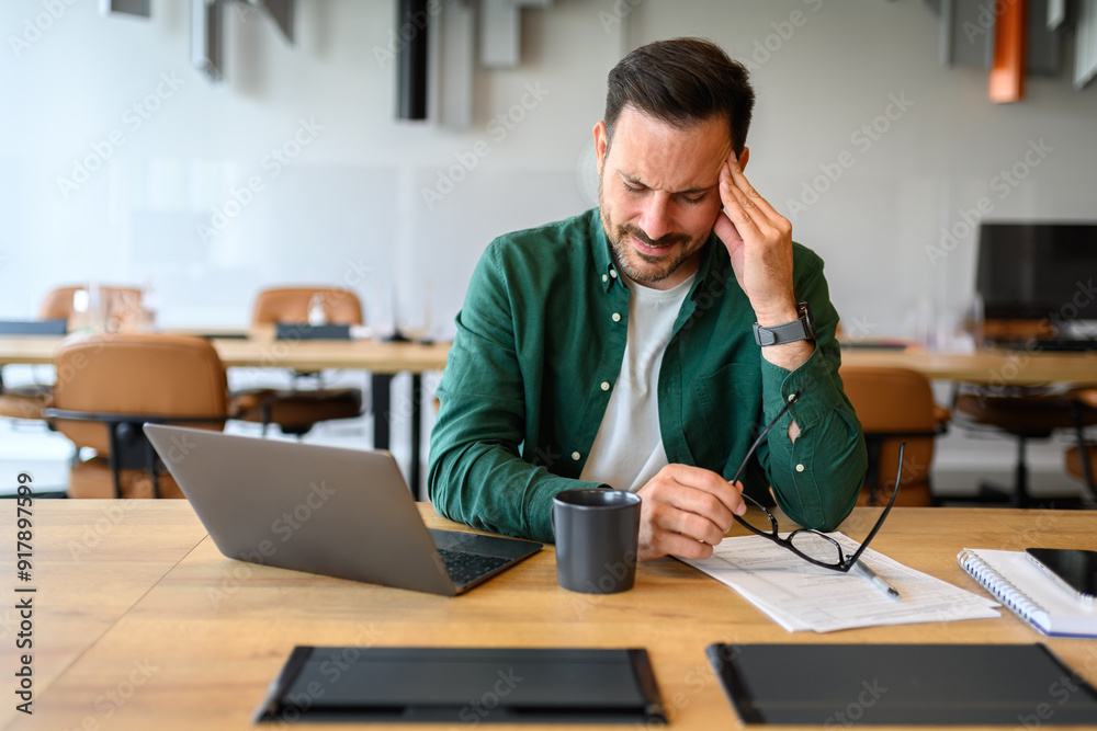 Young businessman holding eyeglasses and touching head in pain while working over laptop in office Young businessman holding eyeglasses and touching head in pain while working over laptop in office