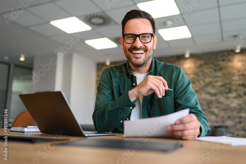 Portrait of handsome entrepreneur with laptop and documents smiling and working at desk in office