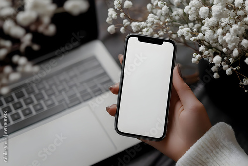 A stylish smartphone displays a blank screen, surrounded by flowers and a laptop, ideal for modern tech and lifestyle concepts. photo