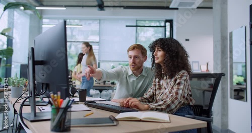 Camera moving closer to two Caucasian colleagues working side by side in modern office. Woman typing on keyboard. Man showing something on notepad. Looking at monitor. Controlling new intern.