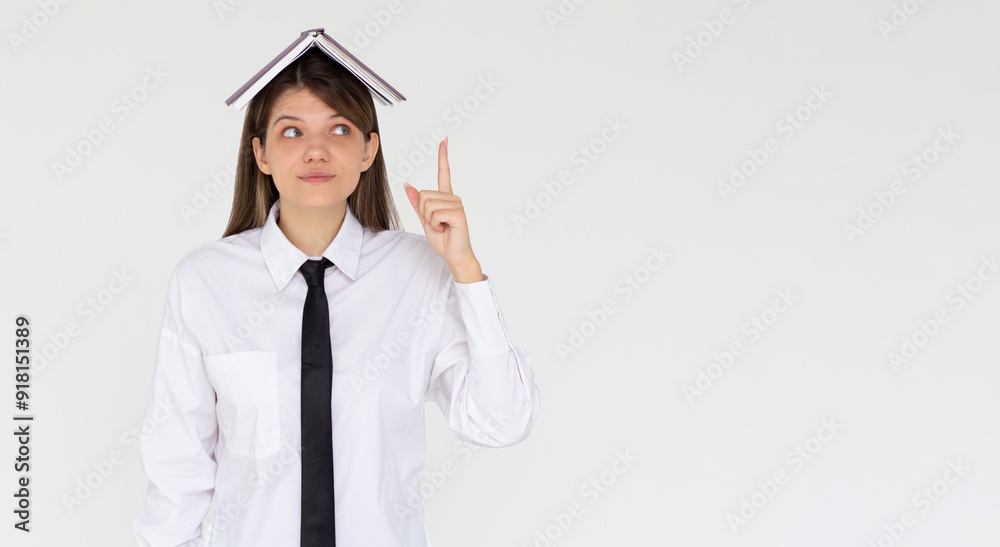 Portrait of funny young smiling student girl with an open book on her head holding her point finger up having an idea. Reading and education concept. Portrait of funny young smiling student girl with an open book on her head holding her point finger up having an idea. Reading and education concept.
