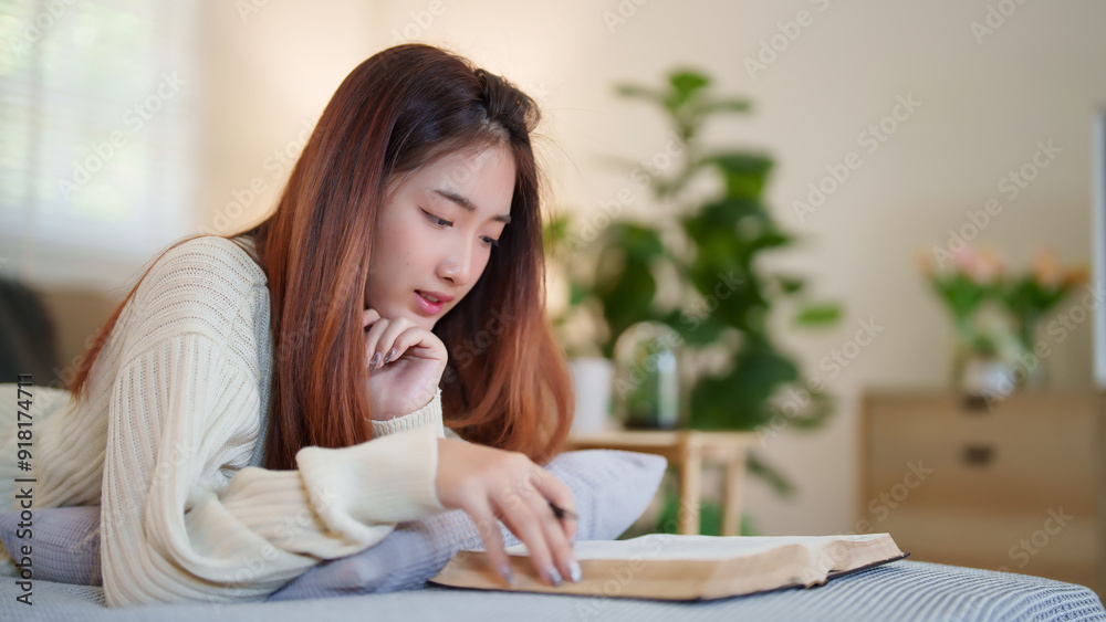 Young asian women in sweater lying on comfortable couch to enjoying with reading literature book and taking notes while relaxation and doing activity to spending time with slow life lifestyle at home. Young asian women in sweater lying on comfortable couch to enjoying with reading literature book and taking notes while relaxation and doing activity to spending time with slow life lifestyle at home.