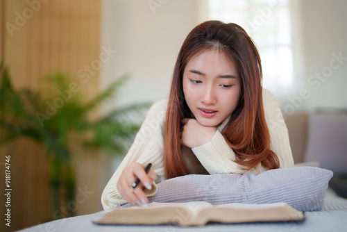 Young asian women in sweater lying on comfortable couch to enjoying with reading literature book and taking notes while relaxation and doing activity to spending time with slow life lifestyle at home.