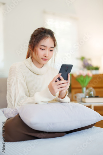 Young asian women in sweater enjoying with surfing entertainment on smartphone while sitting on comfortable couch to relaxation and doing activity for spending time with slow life lifestyle at home