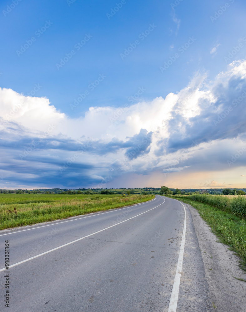 A winding asphalt road stretches out through verdant fields, leading towards a horizon framed by a blue sky dotted with fluffy white clouds. A winding asphalt road stretches out through verdant fields, leading towards a horizon framed by a blue sky dotted with fluffy white clouds.