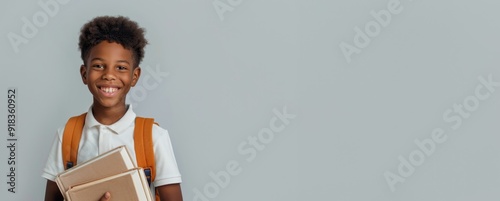 Smiling boy with short curly hair holding books and wearing a white polo shirt with an orange backpack in front of a plain grey background.