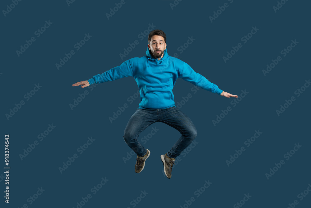 A young man demonstrates his enthusiasm by leaping off the ground in a bright blue hoodie, showcasing his playful spirit and joy. The solid dark background emphasizes his energetic pose A young man demonstrates his enthusiasm by leaping off the ground in a bright blue hoodie, showcasing his playful spirit and joy. The solid dark background emphasizes his energetic pose