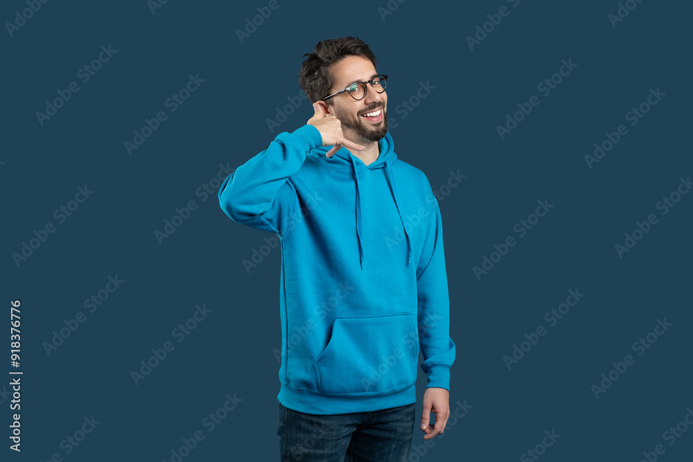 A cheerful guy wearing a bright blue hoodie stands confidently, flashing a warm smile. He makes a playful calling gesture with his hand near his ear A cheerful guy wearing a bright blue hoodie stands confidently, flashing a warm smile. He makes a playful calling gesture with his hand near his ear