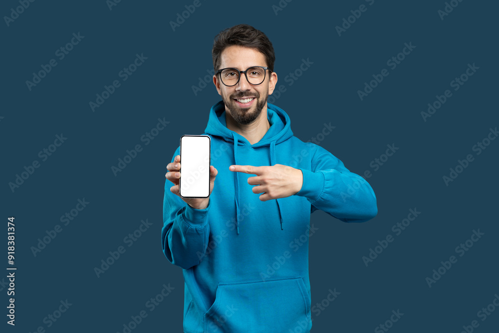 A young man stands against a solid blue background, wearing a blue hoodie and glasses. He is pointing at his smartphone with a cheerful expression, seemingly excited about what he is sharing.