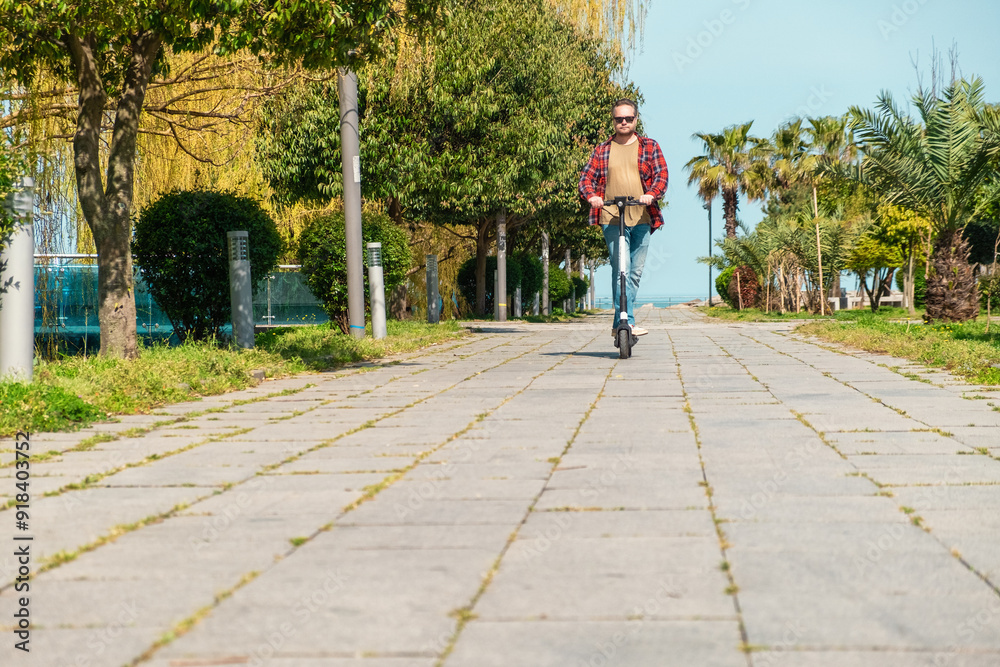 Fototapeta premium Man riding e-scooter on pavement surface in city public park at sunny summer day. Male driving electric scooter outdoors. Eco-friendly city transport and travel. Sustainable lifestyle concept