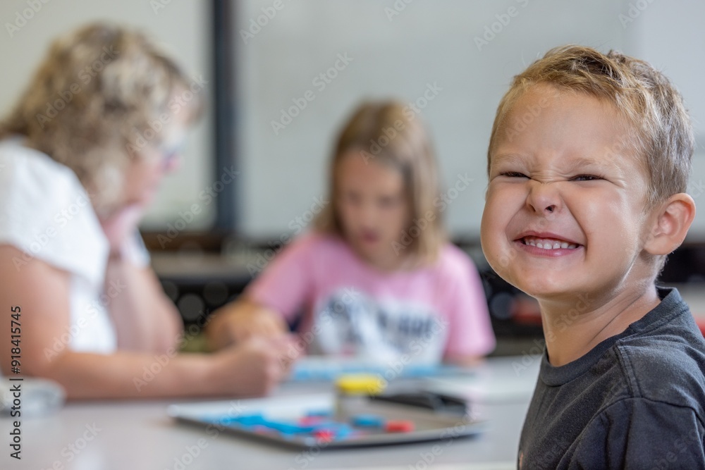 Fototapeta premium A young boy is smiling and sitting at a table with a teacher and a classmate. The scene appears to be a classroom setting, with the boy possibly working on a project or participating in a group activi