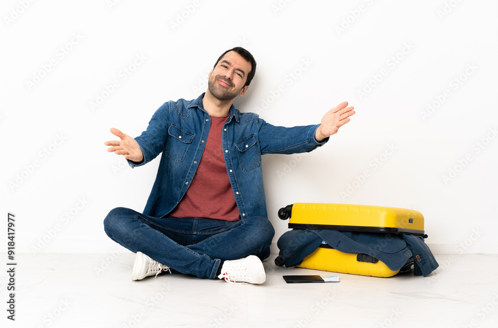 Caucasian handsome man with a suitcase full of clothes sitting on the floor at indoors presenting and inviting to come with hand Caucasian handsome man with a suitcase full of clothes sitting on the floor at indoors presenting and inviting to come with hand