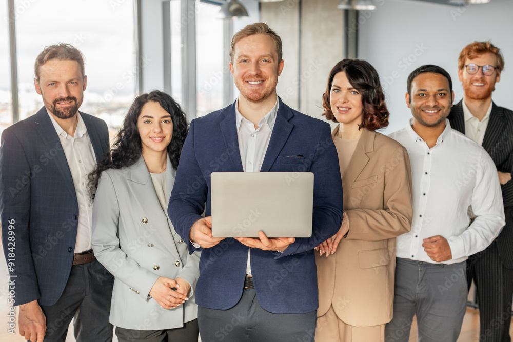 A diverse group of business professionals stand together in a modern office setting. The man in the center is holding a laptop and smiling at the camera, while the others smile and look at the camera A diverse group of business professionals stand together in a modern office setting. The man in the center is holding a laptop and smiling at the camera, while the others smile and look at the camera