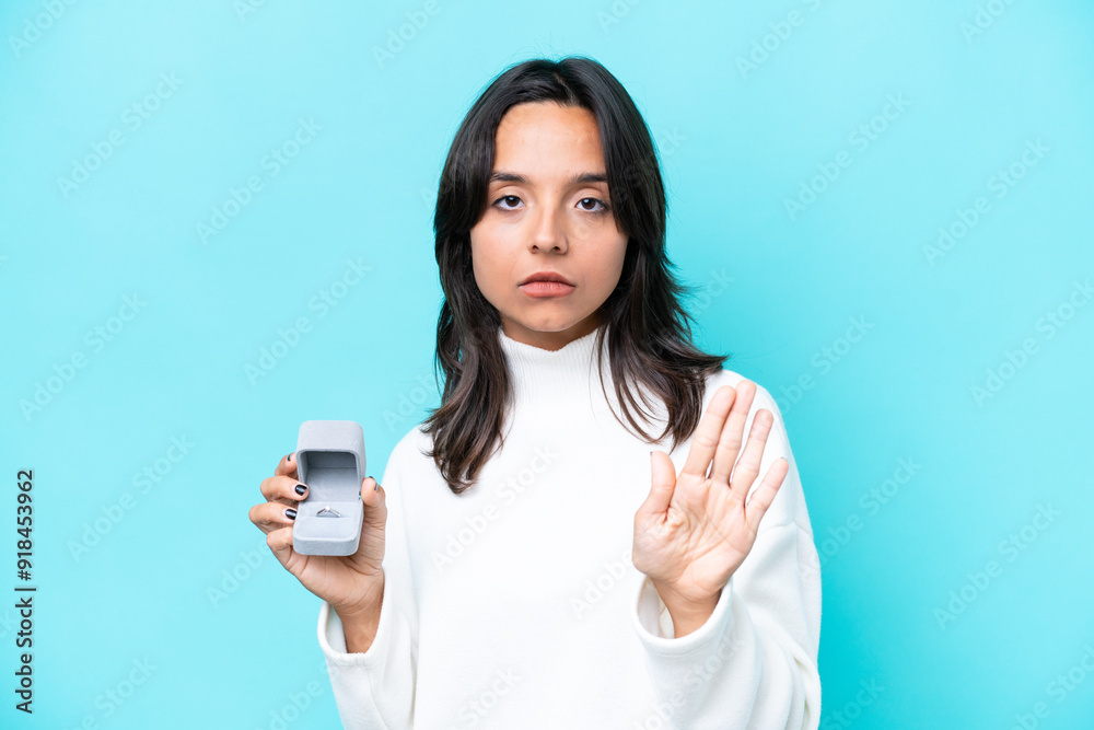 Young hispanic woman holding a engagement ring isolated on blue background making stop gesture Young hispanic woman holding a engagement ring isolated on blue background making stop gesture
