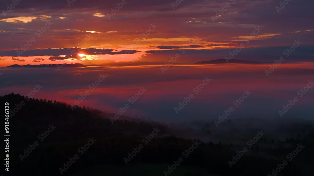 AERIAL: Burnt orange autumn skies gently illuminate the forest-covered hills below the cloudy sky. Spectacular aerial view of the setting sun shining on the dark countryside on an idyllic fall evening