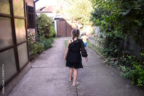 A little girl with two braids in black dress walking through the garden. A little boy with blond hair running in the background. photo