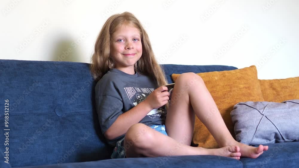 A schoolgirl holds a mobile phone and looks at the smartphone screen. Children's addiction to gadgets. A girl is sitting on the couch at home, an authentic real-time environment A schoolgirl holds a mobile phone and looks at the smartphone screen. Children's addiction to gadgets. A girl is sitting on the couch at home, an authentic real-time environment