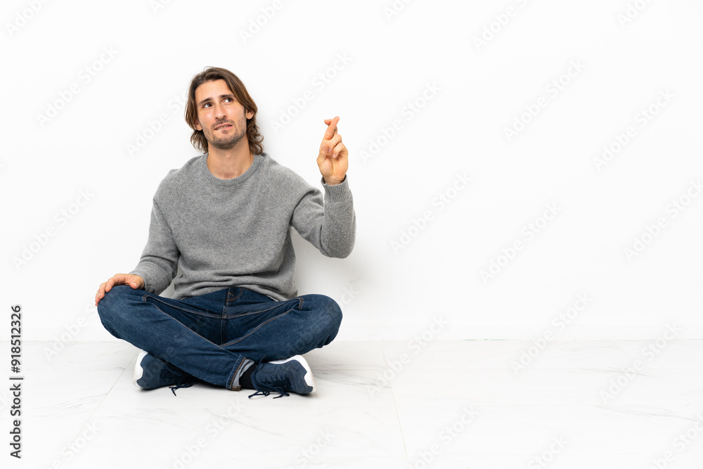Young handsome man sitting on the floor over isolated background with fingers crossing and wishing the best Young handsome man sitting on the floor over isolated background with fingers crossing and wishing the best
