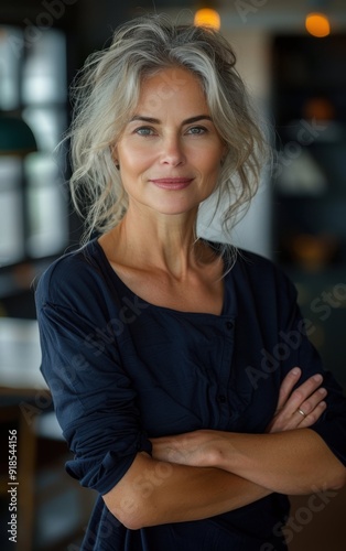 A woman with gray hair is smiling and wearing a blue shirt. She is standing in front of a table with a lamp on it