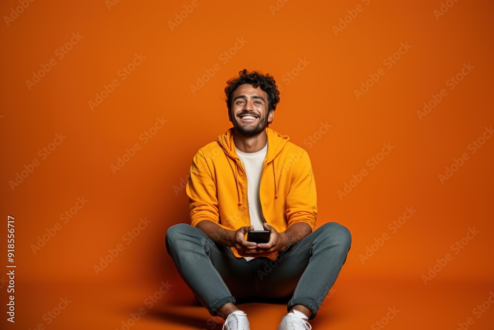 Full body young smiling happy Indian man he wears t-shirt casual clothes sit hold in hand use mobile cell phone look aside isolated on orange red color background studio portrait Full body young smiling happy Indian man he wears t-shirt casual clothes sit hold in hand use mobile cell phone look aside isolated on orange red color background studio portrait