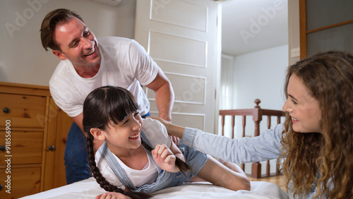Fotografie Happy modern family wakes up their little girl on the weekend with playful tickle expressing their love and affection for their young daughter, laughing and smiling together