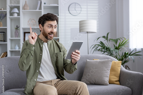 Excited man sitting on sofa using tablet, showing eureka moment with raised finger, concept of discovery or solution. Indoor scene, casual clothing, happy facial expression.