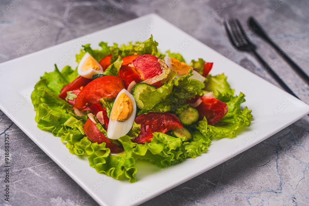 A light vegetable salad for healthy dietary nutrition. Tomatoes, cucumbers and boiled eggs with lettuce and traditional French vinaigrette sauce, on a white plate on a gray table. A light vegetable salad for healthy dietary nutrition. Tomatoes, cucumbers and boiled eggs with lettuce and traditional French vinaigrette sauce, on a white plate on a gray table.