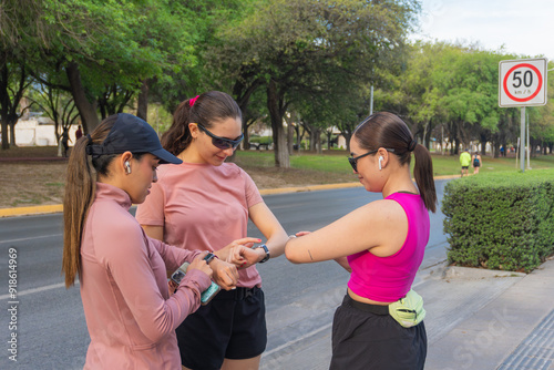 Three latin women in sportswear stand together on the sidewalk, using their fitness trackers during an outdoor workout. Green park and road in the background