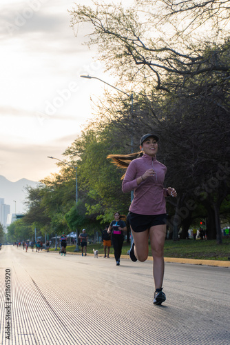 A latin woman in sportswear runs on a city street during an outdoor race. Other runners and trees are visible in the background