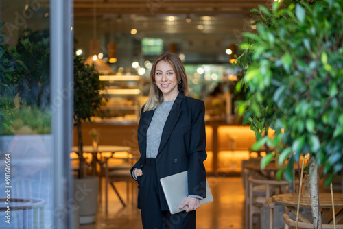 Billede på lærred Confident and smiling businesswoman stands with laptop in doorway of brightly lit cafe, looking at camera