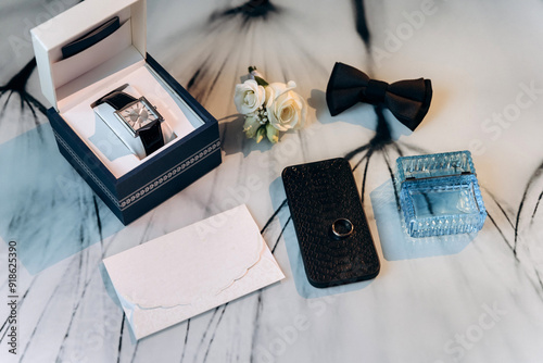 Groom's wedding essentials on table, featuring rings on phone, bow tie, boutonniere, perfume, belt