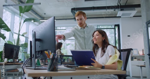 Smart leader standing behind woman and helping her with work at computer. Pretty woman sitting at table and holding folder with documents. Man showing thumbs up to female. Productive teamwork.