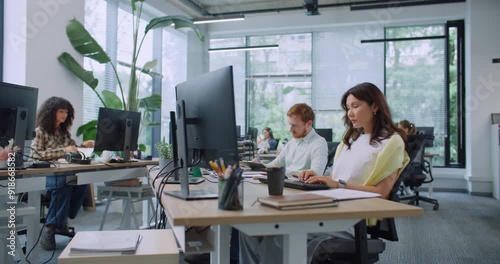 Camera view of busy mixed-raced managers working in large modern office. People sitting at desks with computers. Typing text on keyboards. Entering data on technology devices. Business concept.