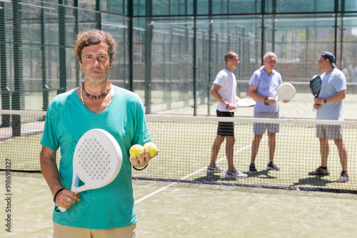 Portrait of positive man standing on padel tennis court, holding racket and ball.