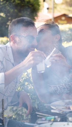 On a summer day at a picnic area, a middle-aged woman kisses her boyfriend on the cheek while clinking their glasses of rakı together photo