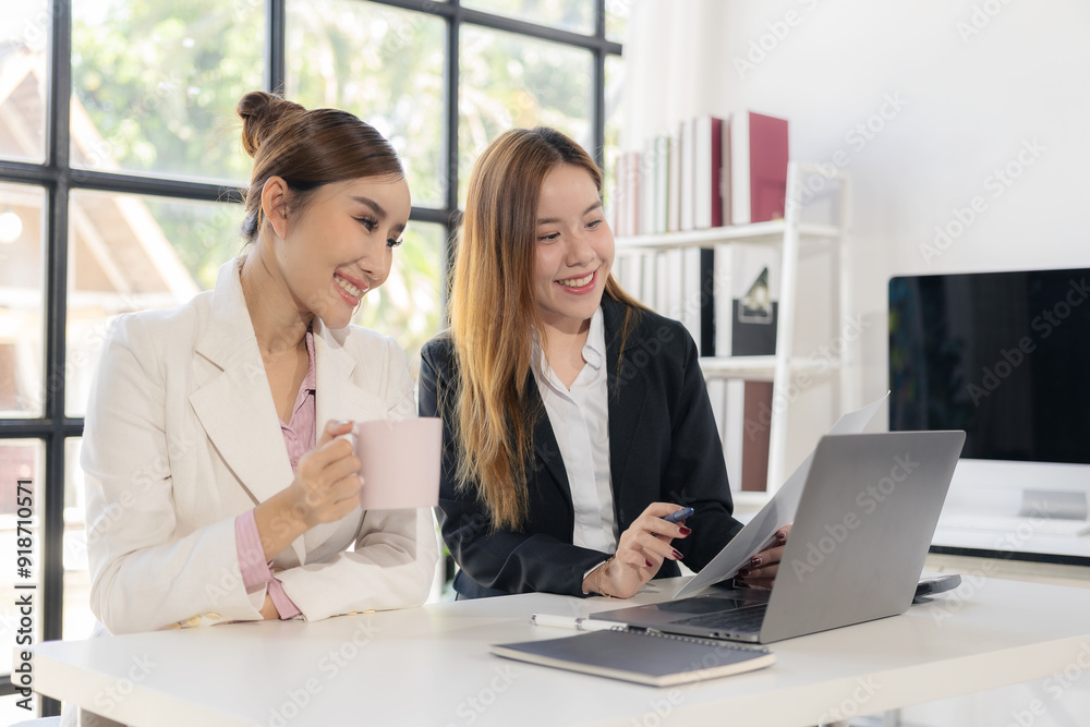 Fototapeta premium Two Asian businesswomen working together, meeting and discussing a new business project, creative team analyzing documents on marketing plan Laptop and documents on the table