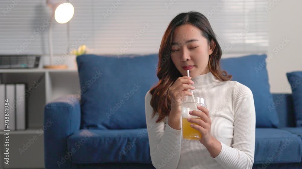 Happy young woman enjoys a refreshing drink at home, smiling and laughing on a cozy blue sofa. She radiates joy and relaxation in her casual indoor setting Happy young woman enjoys a refreshing drink at home, smiling and laughing on a cozy blue sofa. She radiates joy and relaxation in her casual indoor setting