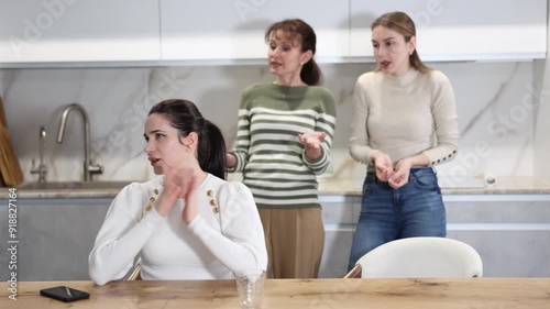 Sad middle-aged woman sitting at table and other women brawling to her standing behind in the kitchen photo