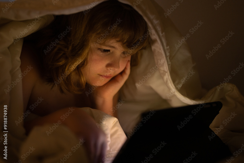 Kid is addicted to tablet, kid use tablet, watching cartoon. Child watching his tablet in the bed. Illuminated child face from device screen. Boy under the covers hold a tablet. Night time. Kid is addicted to tablet, kid use tablet, watching cartoon. Child watching his tablet in the bed. Illuminated child face from device screen. Boy under the covers hold a tablet. Night time.