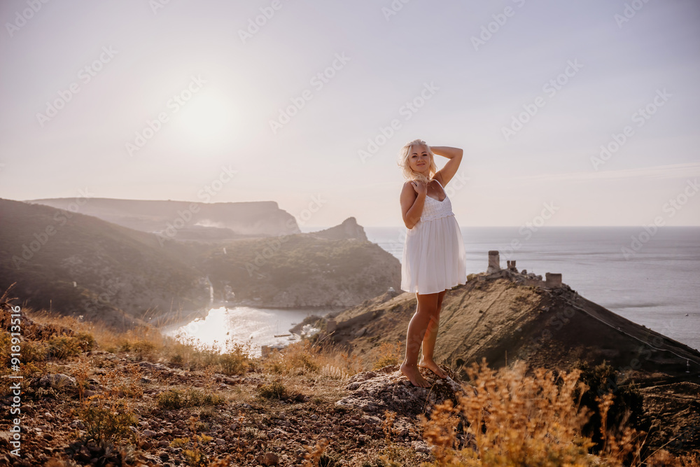 Obraz premium A blonde woman stands on a hill overlooking the ocean. She is wearing a white dress and she is enjoying the view.