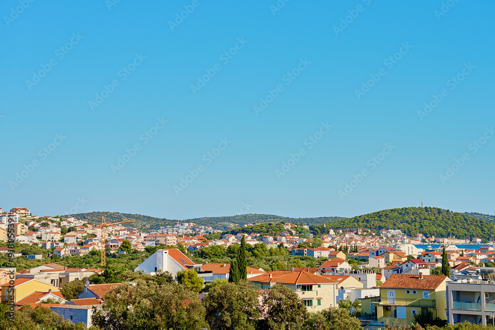 Obraz premium View on coastal town with red roofed houses and lush green hills under clear blue sky. Touristic Mediterranean town near Adriatic sea for summer holidays